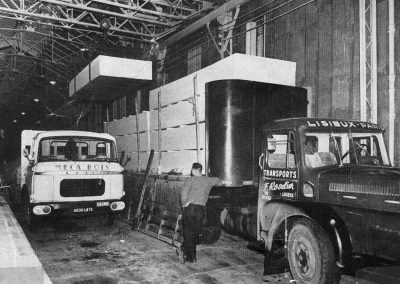 Deux camions chargés de grandes boîtes dans un entrepôt industriel, avec une personne debout entre eux.